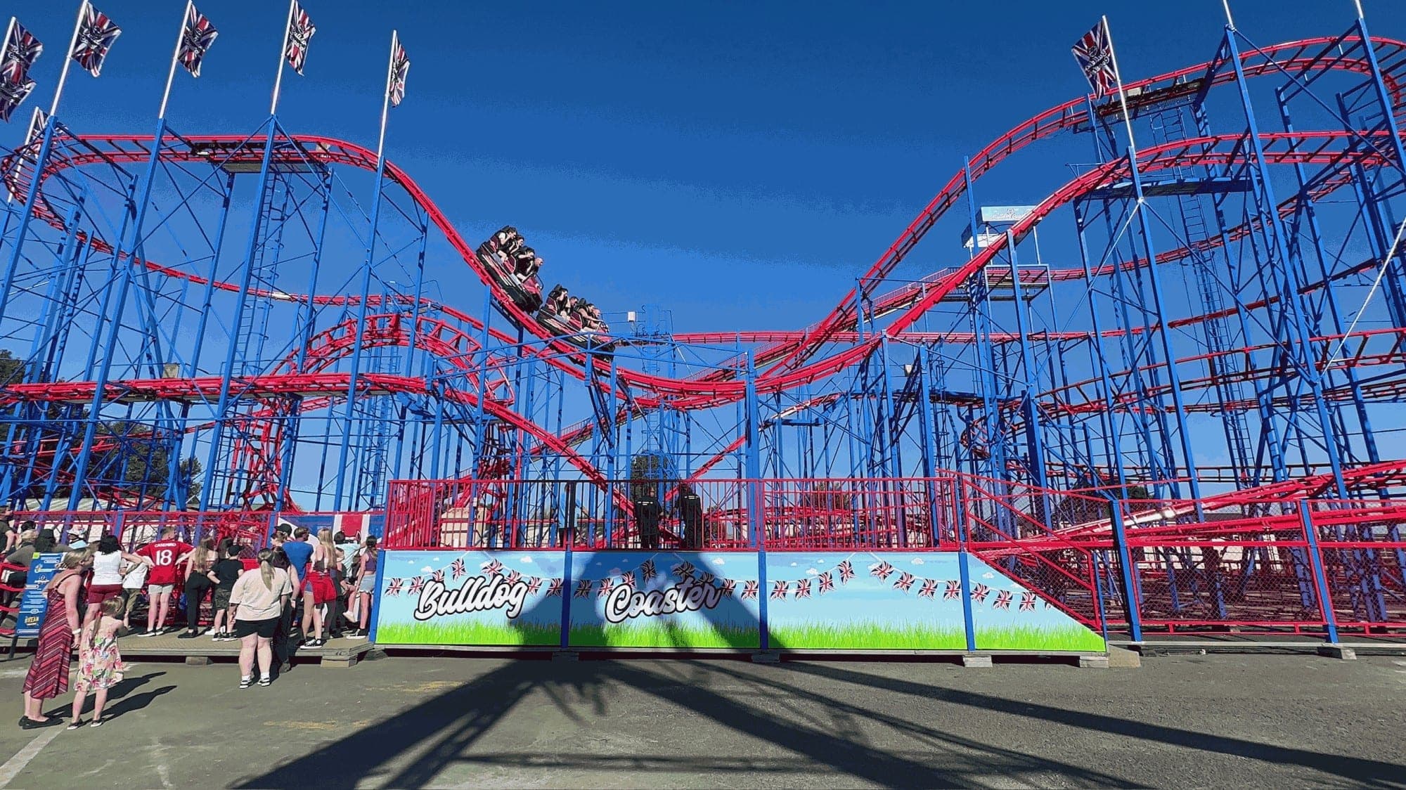 The Bulldog Coaster, red track on blue steel, with its station panels of Union Jack bunting and the ride name in script, and a queue of visitors in front.